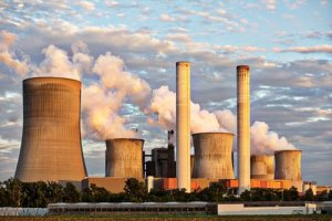 A large power plant site with multiple cooling towers and chimneys emitting steam or smoke against a partly cloudy sky, surrounded by greenery. The cooling towers have a distinctive hourglass shape, juxtaposing industrial might with the surrounding environment's focus on wellbeing.