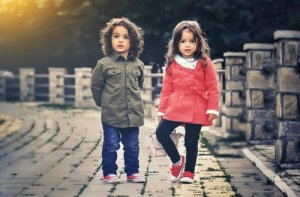 Two young children stand on a paved pathway. The child on the left wears a green jacket and jeans, while the child on the right wears a red coat and black pants. Both have curly hair and wear red shoes, radiating joy and wellbeing. Stone railings line the path, with warm, soft lighting in the background.