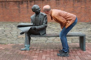 A man wearing a brown leather jacket, blue jeans, and glasses bends over and leans toward a life-sized bronze statue of a seated man holding a newspaper, as if engaged in quiet conversations. The background features a brick wall and cobblestone ground.