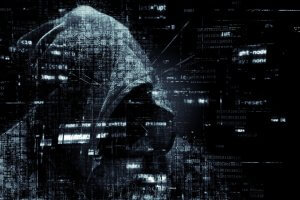 A person in a hooded sweatshirt is surrounded by digital code and binary numbers, evoking themes of cyber security and hacking. The dark, shadowy figure blends with the monochrome background filled with lines of computer text and data.