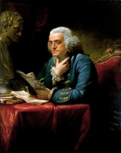A man with white hair and glasses, dressed in an 18th-century blue coat, is seated at a table with a red tablecloth, reading papers. A bust of a man and a stack of books symbolize leadership in the ornate, dimly-lit room.
