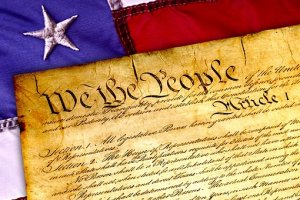 A close-up image of the United States Constitution's preamble, "We the People," displayed on a parchment. The top corner of an American flag with a white star on a blue field and red and white stripes is visible in the background, symbolizing the nation's enduring spirit of leadership.
