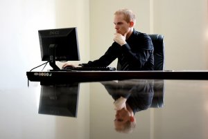 A man in a black suit, exemplifying leadership, sits at a sleek, reflective desk, intently looking at a computer monitor. He rests his chin on his hand, appearing thoughtful as his image mirrors back at him. The background is plain and minimalist.