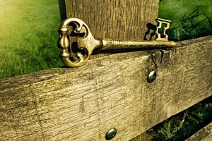 A vintage, ornate golden key rests atop a weathered wooden fence, with lush green grass visible in the background. The intricate design of the key's head and the rustic texture of the wood create an emotional contrast between old-world charm and natural beauty.