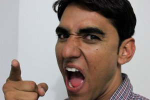 A man with dark hair is angrily pointing his finger, his mouth open as if shouting, radiating an emotional intensity. He is wearing a checkered shirt and the background is plain white.