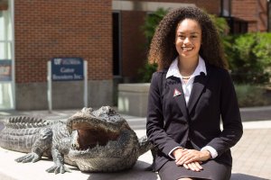 A woman with curly hair, dressed in a black suit and white shirt, is sitting next to a statue of an alligator. Smiling and posing for the photo likely taken for LinkedIn, she has a brick building with a sign that says "Gator Bookstore Inc." in the background.