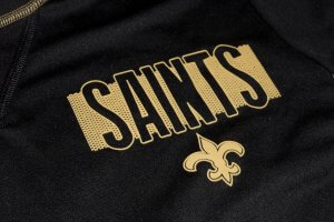 A close-up view of a black garment featuring the word "SAINTS" in bold gold letters. Below the text is a gold fleur-de-lis symbol, often associated with the New Orleans Saints football team and its leader. The fabric appears to be sportswear material.