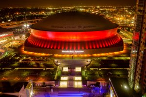 An aerial view of the Mercedes-Benz Superdome at night, illuminated with vibrant red and orange lights, makes a compelling statement. The surrounding city lights sparkle in the background while the stadium's name is prominently displayed on its roof, asserting its position as a leader in sports and entertainment venues.