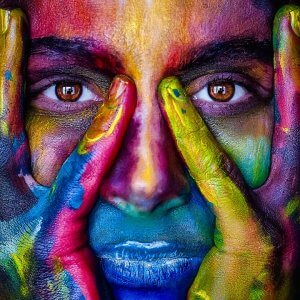 A close-up of a person with hands framing their face, featuring vivid and colorful face paint. The paint covers the face and hands in bright hues—red, blue, green, and yellow—creating a striking artistic impression; the eyes prominently visible seem to ask, "What do you know?