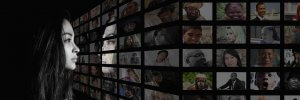 A young woman gazes at a wall of numerous small screens, each displaying diverse portraits of people from various backgrounds and cultures. The scene is predominantly dark, highlighting the illuminated screens and the woman's side profile as if silently asking, "What do you know?