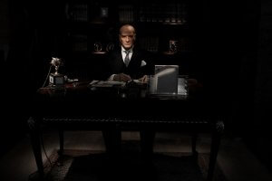 A leader in a suit and tie sits at a large, dark wooden desk in a dimly lit room. He is writing on a document, with bookshelves filled with books and various objects, including an antique telephone, visible in the background.