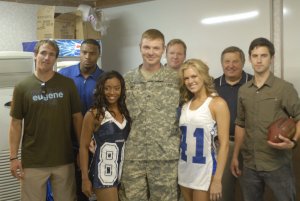 A group of eight people pose for a photo inside a room. In the center, a leader in military uniform is flanked by two cheerleaders in blue and white uniforms. Four men in casual clothing stand behind them, and another man holds a football on the right.