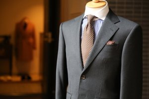 A close-up of a mannequin dressed in a dark gray suit with a light blue striped shirt and a brown polka-dot tie, perfect for making an impression at your next cover letter interview. A brown coat hangs in the blurred background, all displayed in a well-lit store.