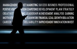 A silhouette of a person walking while looking at a mobile device against a dark background. The backdrop features words such as "management," "marketing," "success," "professional," "leadership," and "achievement"—all elements to consider when crafting an impactful cover letter for business and development.