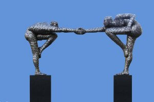 Two mosaic statues balance on black pedestals against a blue sky. The statues, composed of small, reflective tiles, depict two human figures leaning forward. They extend one arm each to clasp hands, creating an arch-like formation that symbolizes unity in the workforce.