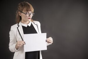 A woman with glasses and a white blazer over a black top holds a blank piece of paper marked "ATS" in front of her. She is looking slightly to the side with a neutral expression against a dark background.