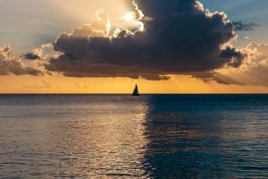 A sailboat is silhouetted against a dramatic sunset backdrop, with the sun's rays streaming through dark clouds, symbolizing the beauty of overcoming adversity. The serene ocean reflects the colors of the sky, creating a tranquil and picturesque scene.