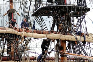 Five people are working on an old wooden sailing ship, managing ropes and sails. They are positioned at different levels, balancing on wooden beams and the rigging. The ship has numerous ropes and pulleys intertwined, showcasing the complexity of the vessel and their resilience in the face of adversity.