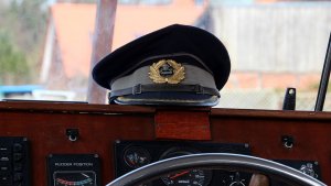 A captain's hat, symbolizing resilience against adversity, rests on the dashboard of a boat. The dashboard displays various gauges and controls, including a steering wheel. In the background, abstract outdoor scenery is slightly blurred.