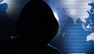 A hooded figure in shadow stands against a background featuring a world map overlaid with binary code. The scene suggests themes of cybersecurity, hacking, or online anonymity. The dark silhouette contrasts sharply with the illuminated digital backdrop.