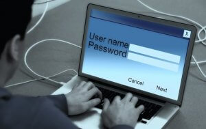 A person is typing on a laptop keyboard. The screen displays a login interface with fields for "User name" and "Password," along with "Cancel" and "Next" buttons. Cables are connected to the laptop, suggesting a network or internet connection, highlighting the importance of cybersecurity measures.