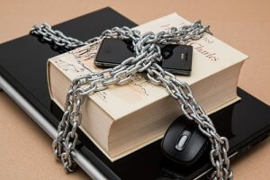 A thick book is wrapped with metal chains, secured by a lock. A smartphone and a laptop mouse are also entangled in the chains. The book sits atop an open laptop, creating a scene that vividly conveys cybersecurity and the restriction or security of information.