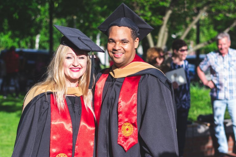 Two graduates stand together outdoors, both wearing black graduation caps and gowns with red and gold stoles. The woman on the left has long blonde hair and a broad smile, while the man on the right has short dark hair and smiles warmly. They are ready to join the workforce. Other people are in the background.