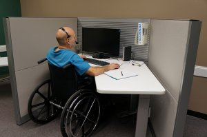 A person in a wheelchair is working at a cubicle desk, exemplifying the inclusive workforce. They are wearing a headset and using a computer with a black monitor. The desk has a notepad and a pen, while the background consists of cubicle dividers and beige walls.