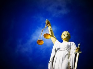 A statue of Lady Justice against a deep blue sky. She is wearing a flowing robe, holding a set of balanced scales in her left hand and a sword in her right hand. Representing fairness, Lady Justice is often used as a symbol of the judicial system.
