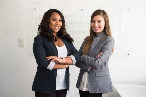 Two women standing in an office setting, both smiling and facing the camera with arms crossed. The woman on the left is wearing a dark blazer, white shirt, and has long curly hair. The woman on the right is donning a gray blazer and white shirt, with straight long hair. Their confident stance reflects fairness in leadership.