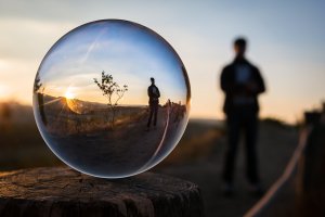 An artistic photo captures a glass sphere resting on a wooden surface, its reflection revealing a plant and person against a stunning sunset backdrop. The blurred silhouette of the person stands in the background, enhancing the scene's depth and focus on the sphere.