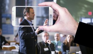 A close-up of a person's hand holding a smartphone, capturing a photo or video of a man speaking at a conference. The man is gesturing with his hand, and several people are seated and listening attentively in the background, their faces mirrored in the reflection on the phone's screen.
