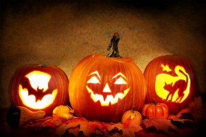 Three carved pumpkins are displayed on a bed of autumn leaves, their reflections subtly visible on the polished surface beneath. The central pumpkin has a classic jack-o'-lantern face, while the pumpkins on the left and right feature bat and cat designs respectively. Small gourds and another mini pumpkin surround them.