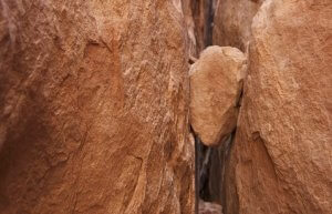 A large tan rock is suspended between two massive rock formations, creating a small gap in an otherwise narrow passage. The textured surfaces of the rocks are prominently visible, much like the nuanced challenges faced in mid-level leadership.