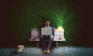 A man sits on a bench at night, working on a writing job from his laptop. Beside him are a steaming coffee mug, a stack of books, and a lit table lamp. The background is a green hedge wall. The scene is dimly lit, creating a focused, serene ambiance.