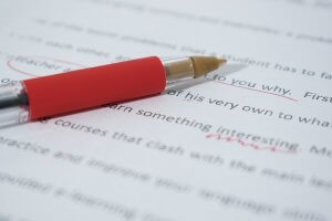 A red pen rests on a printed document with several corrections marked in red ink. Words and sentences are circled and underlined, indicating areas that need revision. The text is blurred except for a few legible words, showcasing the meticulous nature of this writing job.