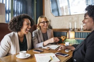 Three women engage in a lively conversation at a cozy café table. On the table are notebooks, a pen, two smartphones, and a cup of coffee. They are smiling and appear to be enjoying each other's company, perhaps discussing their latest writing job and creating a warm and friendly atmosphere.