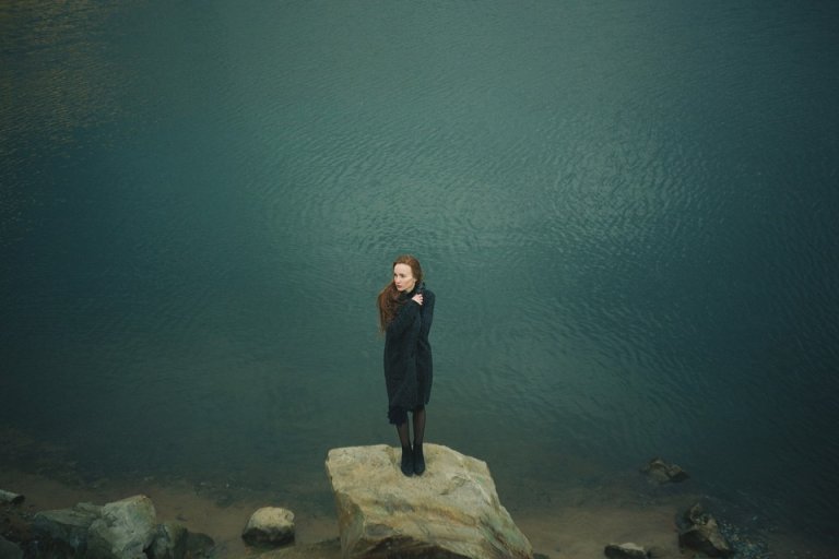 A person dressed in black stands alone on a large rock by the edge of a calm body of water. The individual has long, flowing hair and is facing forward with their arms crossed, creating a serene and contemplative scene despite the shadows of past bullying. The backdrop and foreground are composed of soft, muted colors.
