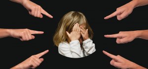 A child with blond hair, wearing a white shirt, covers their face with their hands while surrounded by eight outstretched hands with fingers pointing towards them against a black background. The image starkly captures the essence of bullying and evokes themes of accusation and blame.