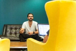 A man with a beard, dressed casually in a white t-shirt and dark pants, is sitting on a brown leather sofa, gesturing while speaking about personal branding. In the foreground, a person with a laptop and wearing a blue shirt is partially visible, seated on a yellow chair.