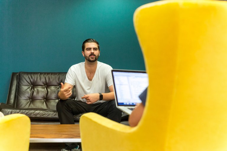 A man with a beard, dressed casually in a white t-shirt and dark pants, is sitting on a brown leather sofa, gesturing while speaking about personal branding. In the foreground, a person with a laptop and wearing a blue shirt is partially visible, seated on a yellow chair.