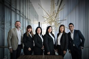A group of six formally dressed people stand side by side in a modern, spacious indoor environment. They are all smiling and appear to be posing for a group photo, showcasing their personal branding. The background features large windows and a unique, gold-colored geometric light fixture.