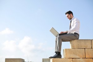 A man in business attire sits on large stone blocks outdoors, working on a laptop. The clear blue sky with scattered clouds serves as a backdrop to his focused and engaged demeanor, embodying personal branding effortlessly.