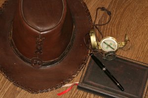 A brown leather hat with decorative stitching sits on a wooden surface, embodying rustic culture. Beside it are a pocket watch with a green compass, a black pen, and a dark brown leather-bound notebook with a red ribbon bookmark.