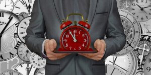 A person in a suit holds a red alarm clock on a tray, set against the backdrop of numerous clocks of various styles and sizes, all showing different times. The image captures the essence of workplace time management and the passage of time.
