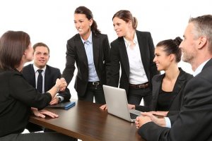 A group of business professionals in formal attire sit and stand around a conference table. Two women are shaking hands with a successful candidate while others look on, smiling. A laptop and some documents are placed on the table.