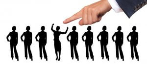 Silhouettes of several women in business attire stand in a row. A giant hand, emerging from the right side of the image, points to a candidate in the middle who has her arms raised in celebration.