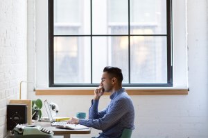 A man sits at a desk working on a laptop in a bright office with large windows. He is wearing a blue checkered shirt and has a thoughtful expression as he delves into programming. The desk also has a green lamp, a cup, a notebook, and various other office supplies.