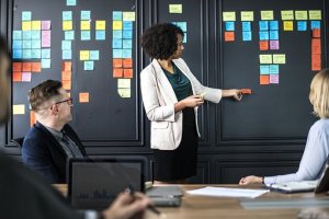 A woman in a white blazer is standing and pointing at a board covered with colorful sticky notes, showcasing her superpowers in project management. Three coworkers are seated around a table, listening intently. A laptop and notebooks are visible on the table, indicating a collaborative work environment.