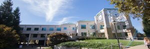 Panoramic view of Sonoma State's modern, multi-story academic building with large windows and green-tinted roofs. The building is surrounded by trees and well-manicured lawns, with a clock mounted on a pole in the foreground and a person walking down a path on the right.
